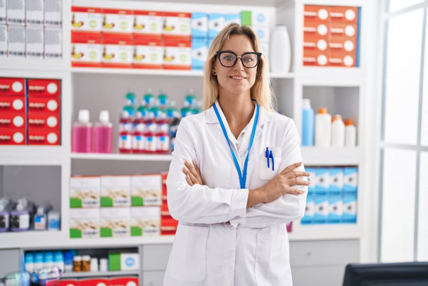 Joven mujer rubia farmacéutica sonriendo confiada de pie con los brazos cruzados gesto en la farmacia - Foto, imagen