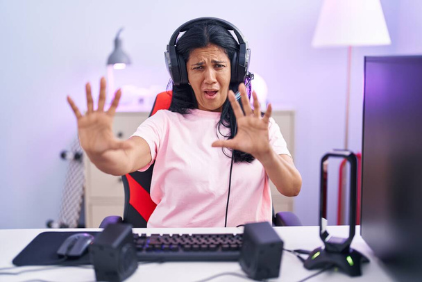 Зріла іспаномовна жінка, яка грає в відеоігри вдома, боїться і налякана виразом страху, зупиняє жест руками, кричить у шоці. панічна концепція
.  - Фото, зображення