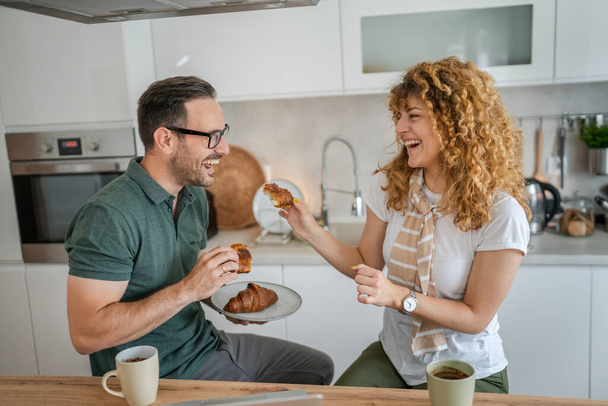 Kaukasisch paar man en vrouw ontbijten in de keuken eten croissant en koffie dagelijks ochtend routine huishoudelijk gezinsleven echte mensen kopiëren ruimte - Foto, afbeelding
