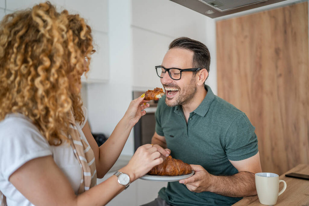 Caucasian couple man and woman having breakfast in the kitchen eat croissant and coffee daily morning routine domestic family life real people copy space - Zdjęcie, obraz