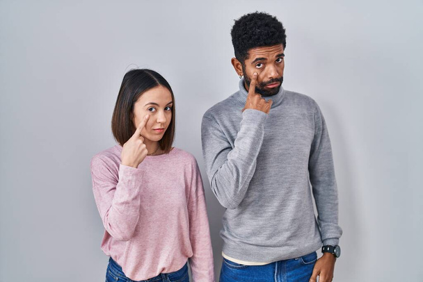Young hispanic couple standing together pointing to the eye watching you gesture, suspicious expression  - 写真・画像