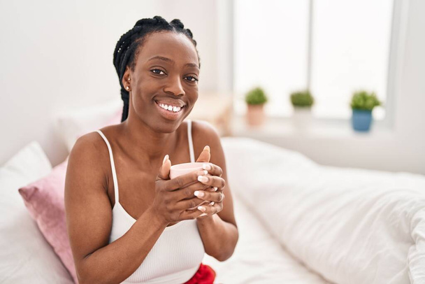 Αφροαμερικανή γυναίκα που πίνει καφέ καθισμένη στο κρεβάτι στο υπνοδωμάτιο - Φωτογραφία, εικόνα