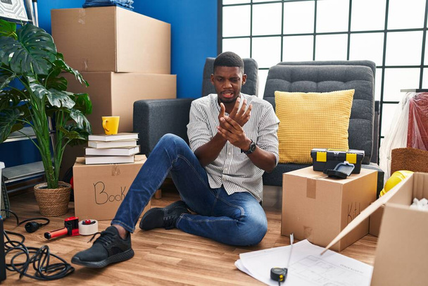 Uomo afroamericano seduto sul pavimento in una nuova casa che soffre dolore alle mani e alle dita, infiammazione dell'artrite  - Foto, immagini