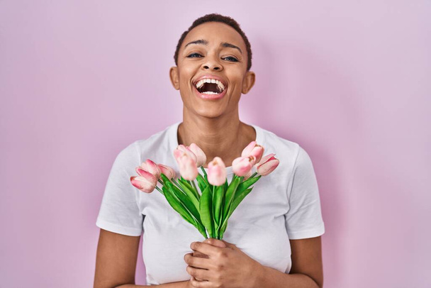 Gyönyörű afro-amerikai nő kezében csokor rózsaszín tulipán virágok mosolyogva és nevetve hangosan ki, mert vicces őrült vicc.  - Fotó, kép