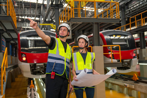 Két profi mérnök vagy technikus munkások férfi és nő megvitatni rajz papírt, és a férfi pont a bal oldalon is marad az elektromos vonat előtt a gyárban munkahelyi vagy karbantartási központ. - Fotó, kép