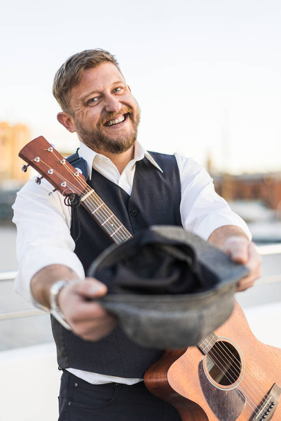 A smiling street musician looking at camera and carrying his guitar while asking for tips - Fotografie, Obrázek