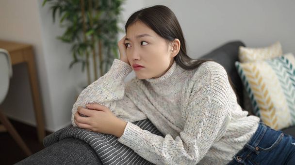 Joven mujer china sentada en un sofá con expresión seria en casa - Foto, Imagen