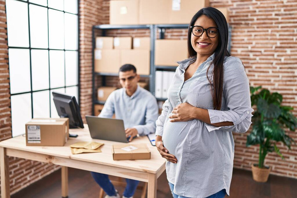 Young hispanic woman expecting a baby working at small business ecommerce looking positive and happy standing and smiling with a confident smile showing teeth  - Photo, Image