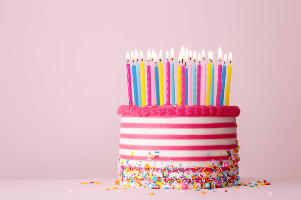 Pembe ve beyaz çizgili tereyağı ve pembe arka planda bir sürü doğum günü mumuyla süslenmiş doğum günü pastası. - Fotoğraf, Görsel