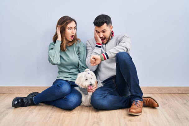 Молодая латиноамериканская пара, сидящая на полу с собакой, глядя на часы, беспокоится, боится опоздать  - Фото, изображение