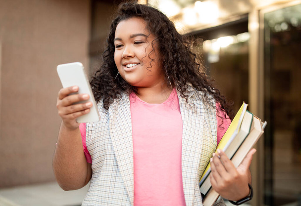 Boldog túlsúlyos, vegyes diáklány, aki mobilon SMS-ezik, és könyveket tart a modern egyetemi épület előtt pózolva. Portré vidám tanuló okostelefon segítségével app online tanulmány - Fotó, kép