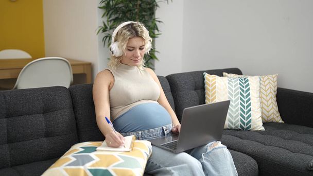 Young pregnant woman using laptop and headphones writing on notebook studying at home - Zdjęcie, obraz