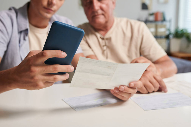 Young man helping grandfather to pay utility bills via mobile application - Photo, Image
