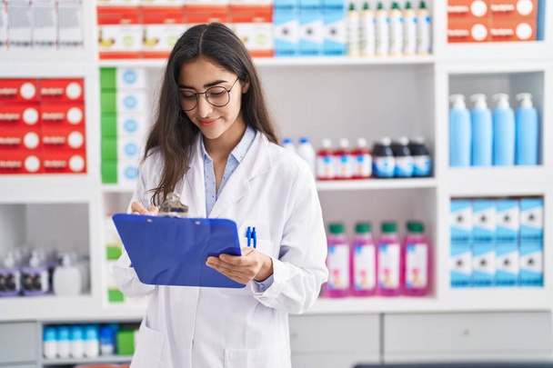 Young hispanic girl pharmacist writing on report clipboard at pharmacy - Foto, afbeelding