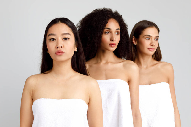 Νεαρές όμορφες πολυφυλετικές γυναίκες ποζάρουν τυλιγμένες σε λευκές πετσέτες μπάνιου, Ασιάτισσα κυρία κοιτάζοντας την κάμερα, τρία μοντέλα ποζάρουν μαζί σε γκρι φόντο - Φωτογραφία, εικόνα