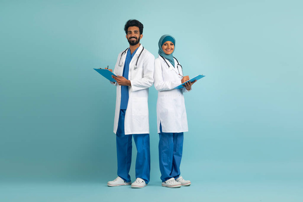 Arabische man en vrouwelijke arts in uniform poseren over Blue Studio achtergrond, professionele moslim medische werkers staan met klembord in handen, klaar voor behandeling, volledige lengte schot met kopieerruimte - Foto, afbeelding