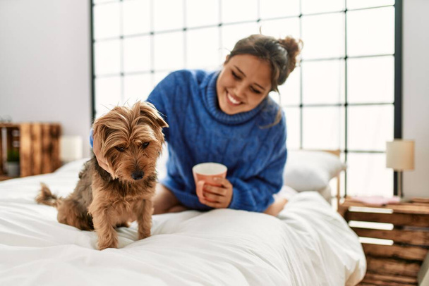 Young beautiful hispanic woman drinking cup of coffee sitting on bed with dog at bedroom - Фото, зображення