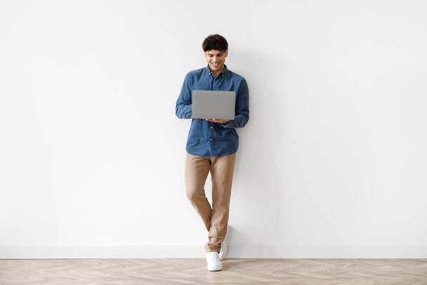 Интернет и работа. Счастливый ближневосточный фрилансер, использующий ноутбук, стоящий на белой стене в помещении. Молодой человек, просматривающий веб на портативном компьютере дома. Полноразмерный снимок, свободное пространство - Фото, изображение