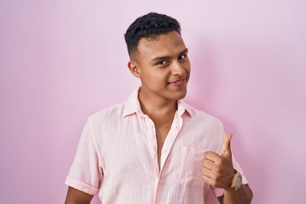 Young hispanic man standing over pink background doing happy thumbs up gesture with hand. approving expression looking at the camera showing success.  - Foto, Imagem