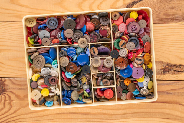 Many buttons in a plastic container on a wooden table, macro, top view. - Фото, зображення