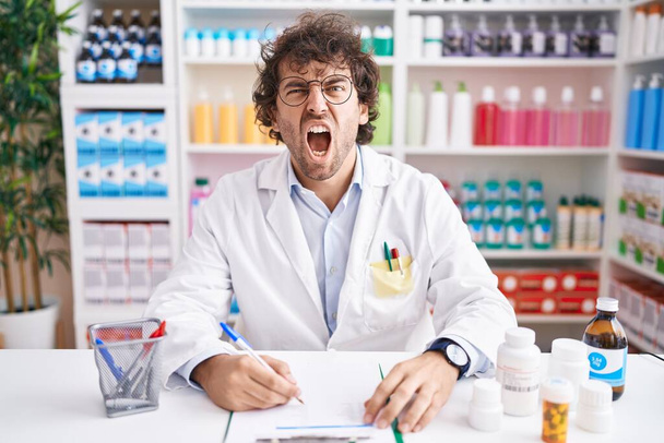 Hispanic young man working at pharmacy drugstore angry and mad screaming frustrated and furious, shouting with anger. rage and aggressive concept.  - Photo, Image