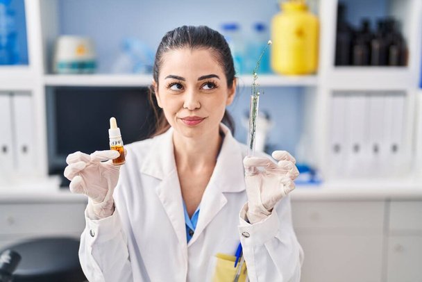 Młoda kobieta robi ekstrakcję zioła w laboratorium uśmiechając się patrząc w bok i patrząc myśląc.  - Zdjęcie, obraz