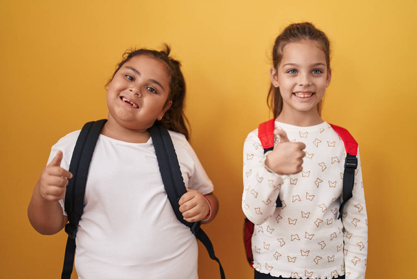 Deux petites filles amies portant un sac à dos étudiant souriant heureux et positif, pouce levé faire excellent et signe d'approbation  - Photo, image