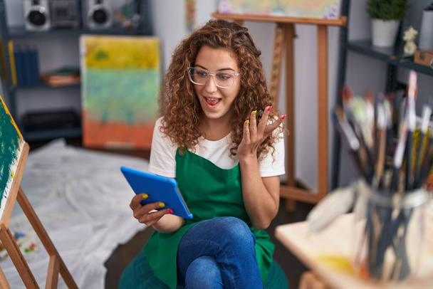 Young caucasian woman painter sitting at art studio doing video call with tablet screaming proud, celebrating victory and success very excited with raised arm  - Photo, Image