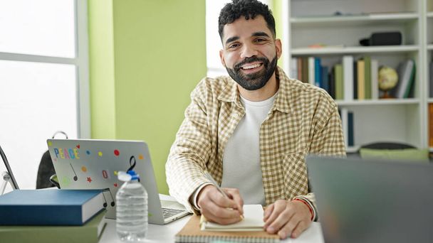 Young hispanic man student using laptop writing notes at library university - Фото, зображення