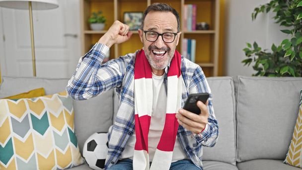Mann mittleren Alters unterstützt Fußballmannschaft zu Hause mit Smartphone - Foto, Bild