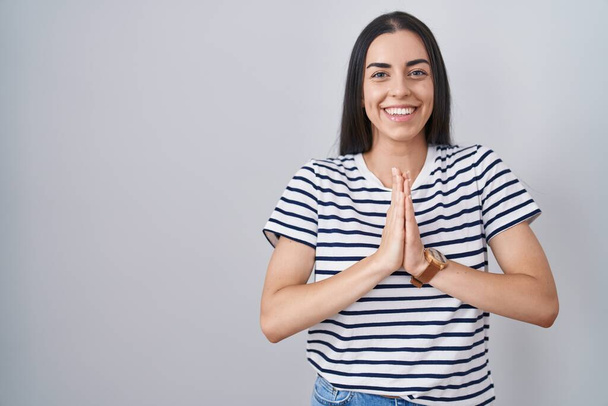 Młoda brunetka kobieta w pasiastej koszuli modli się dłońmi razem prosząc o przebaczenie uśmiechając się pewnie.  - Zdjęcie, obraz