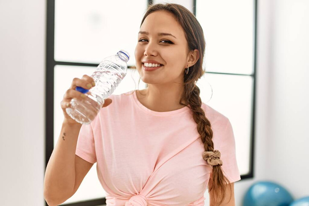 Νεαρή όμορφη ισπανόφωνη γυναίκα χαμογελά με αυτοπεποίθηση πόσιμο νερό στο αθλητικό κέντρο - Φωτογραφία, εικόνα