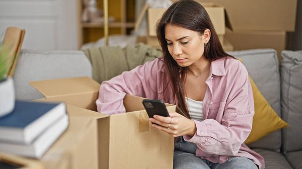 Молодая красивая латиноамериканка с помощью смартфона распаковывает картонную коробку в новом доме - Фото, изображение