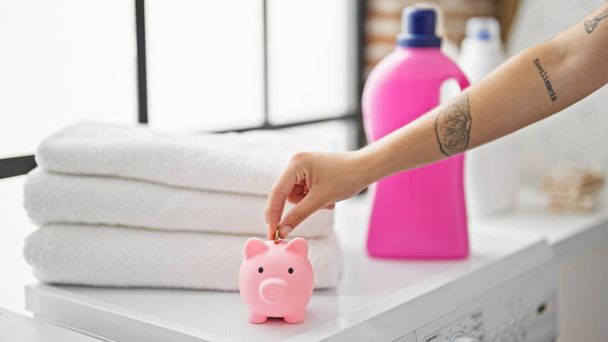 Young beautiful hispanic woman inserting coin on piggy bank at laundry room - Photo, Image