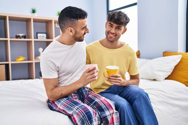 Two man couple drinking coffee sitting on bed at bedroom - Foto, imagen