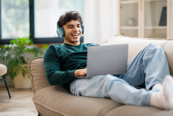 Онлайн веселье. Улыбающийся арабский парень в беспроводных наушниках с ноутбуком дома, расслабляющийся на диване, смотрящий фильм на компьютере или слушающий музыку. Фрилансер с удаленным доступом - Фото, изображение