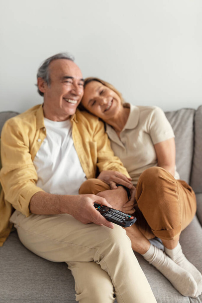Happy elderly senior couple hugging, sitting on sofa and enjoy free time together, watch tv, selective focus on remote controller. Rest at home, love and relationships - Photo, Image