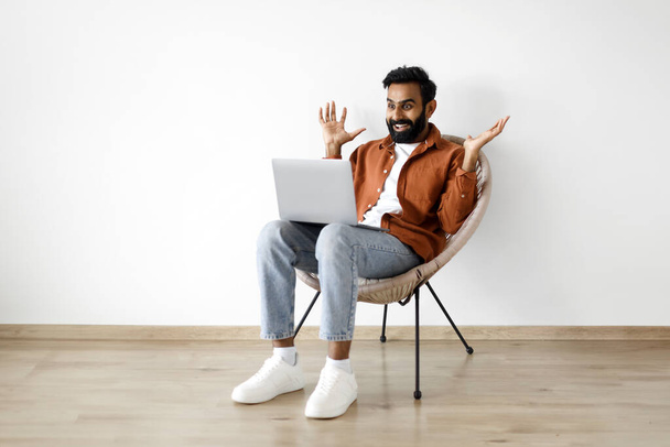 Wow online offer. Эмоциональный ближневосточный мужчина, использующий ноутбук и кричащий от радости, празднуя новости, сидя на уютном стуле и раскладывая руки в помещении. Успешная концепция карьеры фрилансера - Фото, изображение