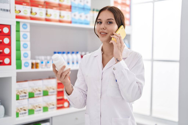 Young woman pharmacist talking on smartphone holding pills at pharmacy - Foto, Bild