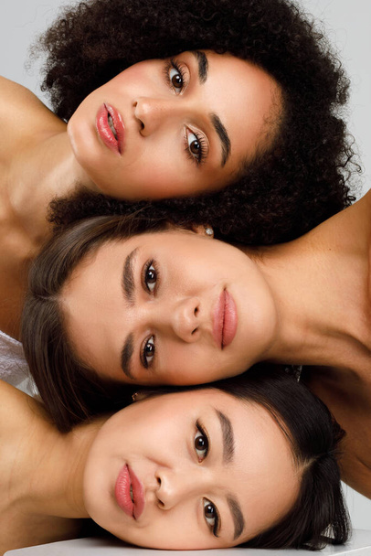 Natural diverse beauty. Portrait of young three multiethnic ladies with perfect face skin and natural makeup posing over gray studio background, vertical panorama shot - Fotó, kép