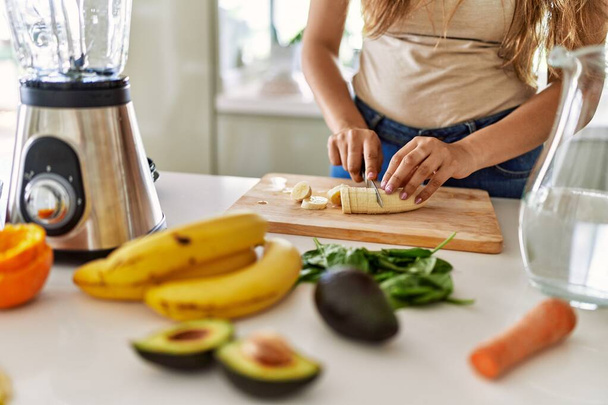 Young beautiful hispanic woman preparing vegetable smoothie with blender cutting banana at the kitchen - Foto, imagen