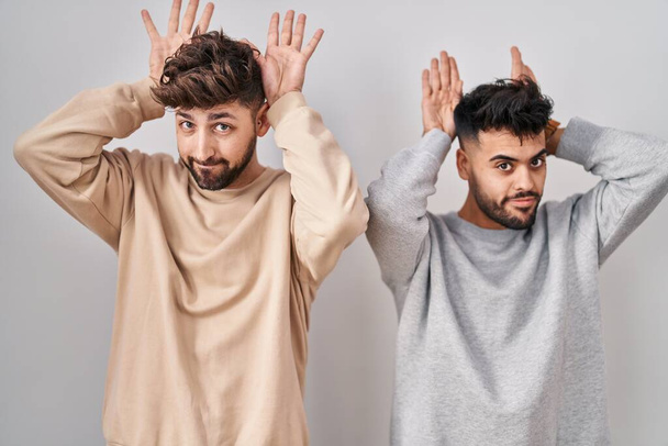 Young homosexual couple standing over white background doing bunny ears gesture with hands palms looking cynical and skeptical. easter rabbit concept.  - Photo, Image