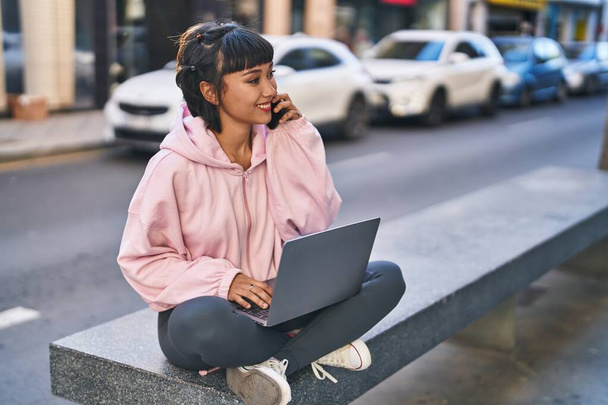 Νεαρή γυναίκα που χρησιμοποιεί φορητό υπολογιστή μιλώντας στο smartphone κάθεται στον πάγκο στο δρόμο - Φωτογραφία, εικόνα