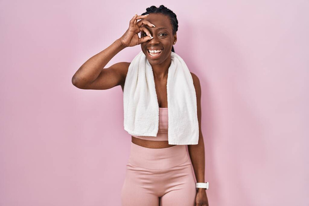 Красивая черная женщина в спортивной одежде и полотенце на розовом фоне делает хорошо жест с улыбкой на руке, глаза смотрят сквозь пальцы с счастливым лицом.  - Фото, изображение