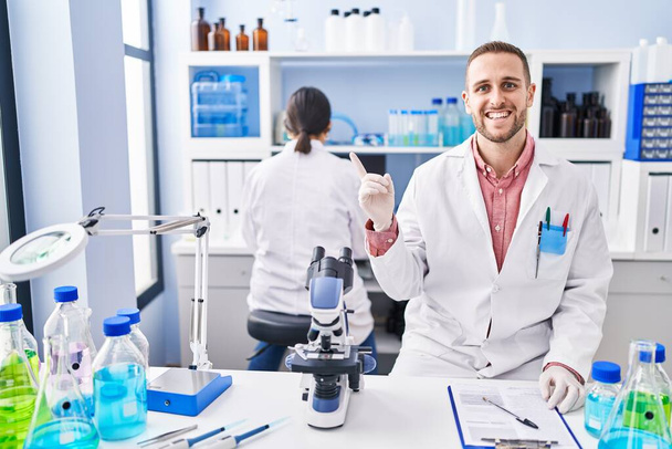 Młody człowiek pracujący w laboratorium naukowca uśmiechający się z uśmiechem na dłoni i palcu.  - Zdjęcie, obraz