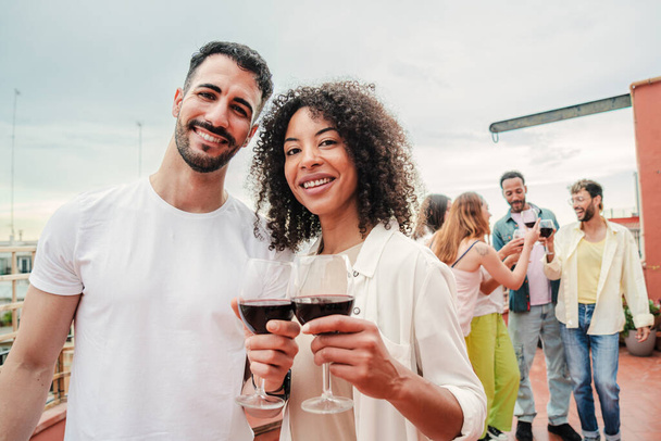 On foreground a multiracial couple toasting the wine glasses, smiling and looking at camera together, at background their friends clinking on rooftoop a party celebration. Patio social reunion at home - Foto, Bild