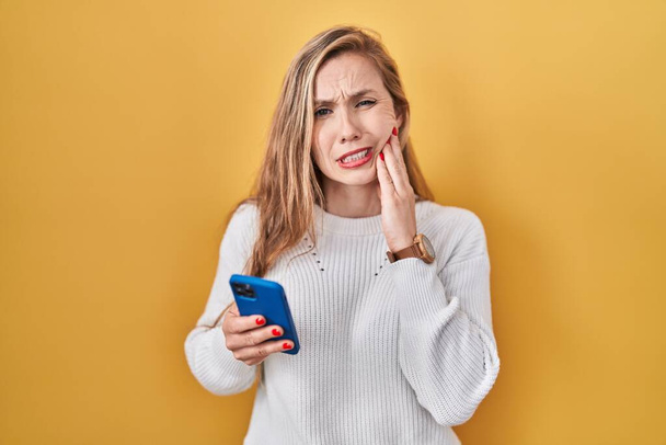 Young blonde woman using smartphone typing message touching mouth with hand with painful expression because of toothache or dental illness on teeth. dentist concept.  - Foto, Bild