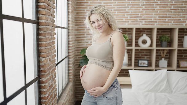 Young pregnant woman touching belly standing at bedroom - Zdjęcie, obraz