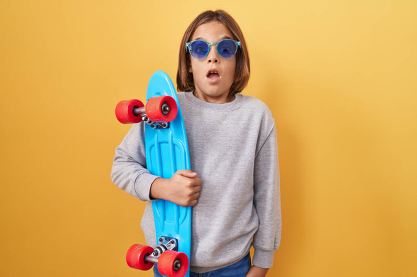 Little hispanic boy wearing sunglasses holding skate scared and amazed with open mouth for surprise, disbelief face  - Foto, Imagem
