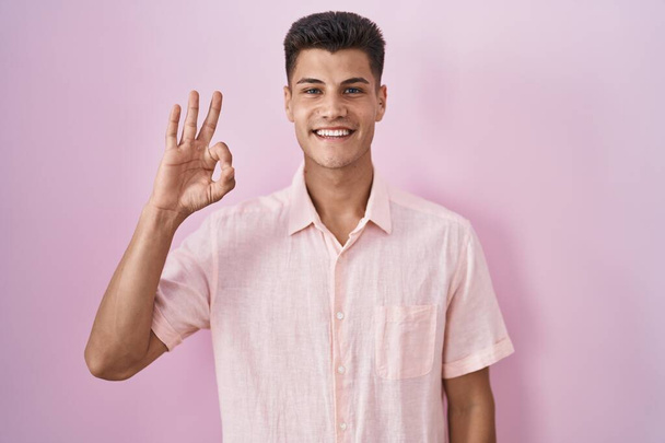ピンクの背景の上に立つ若いヒスパニック系の男が手と指でOKサインをして積極的に笑顔。成功した表現.  - 写真・画像